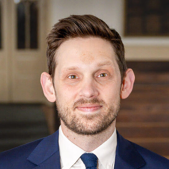 Rabbi Nathan Kamesar, standing in the sanctuary. Rabbi Kamesar is a white man with brown hair and a short beard / mustache. He is wearing a blue suit and tie and smiling into the camera.